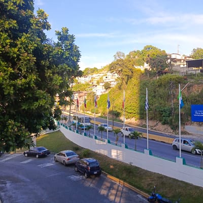 The image depicts an elevated view of a roadway with cars and a bus moving along it, situated in Carrizal, Venezuela. The scene is framed by lush green trees on the left and a hillside covered in residences on the right. The sky is clear blue with a few wispy clouds.
In the foreground, a dark sedan and a silver sedan are parked on a paved area. A dark blue hatchback is parked on the side of the road, partially on the grass. A blue motorcycle lies on its side on the grass near the edge of the paved area.
The roadway itself is lined with white barriers and decorative green posts. Several flags are erected along the roadway, including what appears to be the Venezuelan flag, and flags with blue and white stripes. Palm trees are planted along the median.
Traffic on the road includes a white and blue bus, several cars, and at least one white SUV. The houses on the hillside are of varying sizes and colors, predominantly white, and are built into the steep terrain.
In the distance, a cell tower is visible atop the highest hill. A large billboard is also present on the right side of the image with text that appears to read "La mejor fibra de Venezuela." The overall impression is of a vibrant, urban landscape set against a natural, hilly backdrop under a bright, sunny sky.