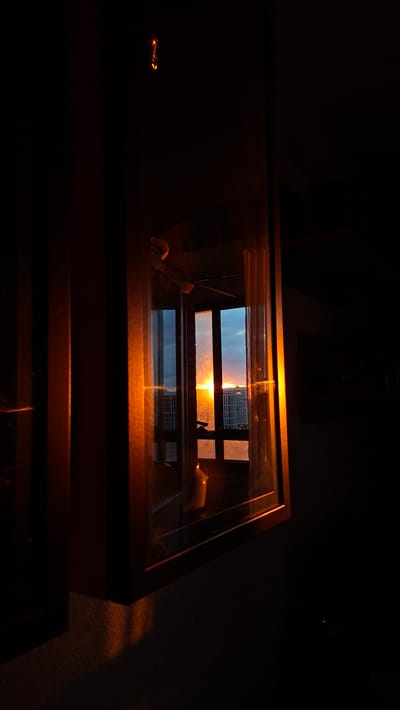 The image is a dimly lit indoor shot with an orange glow from a sunset visible through a window. The window has multiple panes and shows a cityscape with tall buildings in the distance. The sunset is bright and casts a warm light on the interior, illuminating a portion of the window frame and a sheer curtain. The overall scene suggests an evening view from an apartment or building in a city, possibly Russia, given the reference to Unknown, Russia, though no specific landmarks confirm this. The image is dark, with the exception of the light from the sunset and interior lighting reflecting on surfaces. There are no people or animals visible in the frame.