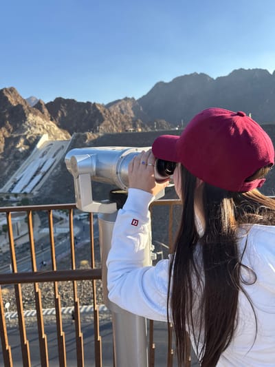 A female subject, viewed from behind and slightly to the right, is looking through a silver coin-operated binocular viewer mounted on a stand. She wears a dark red baseball cap, a white long-sleeved shirt with a red square logo on the left cuff, and rings on her right hand. Her hands are positioned on the viewer's eyepieces. The viewer is situated on an observation deck with a dark brown metal railing in the immediate foreground. In the background, rugged, arid, brown mountains rise under a clear blue sky. A large, sloped concrete structure, resembling a dam or spillway, is visible on the left side of the mountainside. Below the observation point, roads and infrastructure are visible winding through the mountainous terrain. The scene is located at a mountain viewpoint in Hatta, United Arab Emirates, likely overlooking the Hatta Dam area.