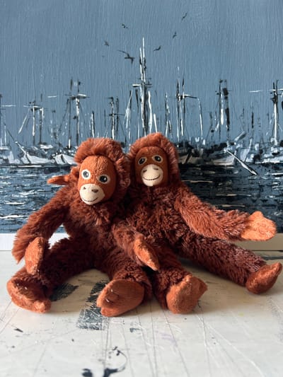Two brown plush orangutans are positioned on a surface with a painting in the background. The orangutans are lying down, with their arms and legs spread out. One orangutan is positioned slightly above and behind the other. Both have round faces with stitched eyes and smiling mouths. The one in front has its head turned to the side, while the one behind is looking forward. The background is an abstract painting with vertical and horizontal brushstrokes in shades of blue, black, and white, giving the impression of a cityscape or a textured wall. The surface beneath the orangutans is a lighter color and appears to be a table or floor with some faint markings. The lighting suggests it is daytime, and there are no indications of weather conditions. The overall mood is playful and whimsical due to the presence of the stuffed animals. The location context of Mollet del Vallès, Spain, does not provide any specific visual cues within the image itself. There is no visible text in the image.