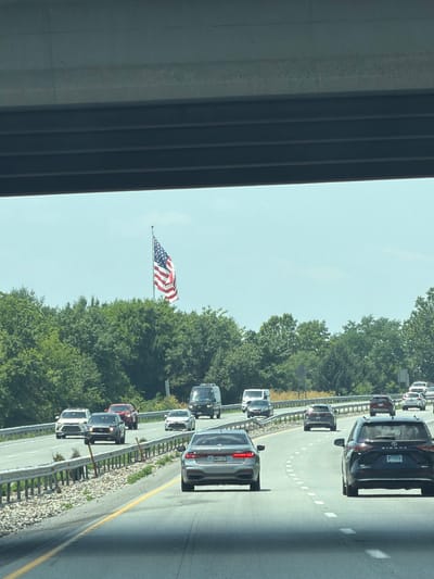 A wide shot captures an American flag flying from a flagpole, positioned behind a line of trees. The flag is seen against a bright, hazy sky. In the foreground, a multilane highway is filled with traffic, with several cars and SUVs visible. A concrete overpass looms in the upper portion of the frame, casting a shadow over the highway. The environment appears to be a semi-rural or suburban setting, with dense green trees bordering the highway. The road surface is asphalt, with distinct lane markings. The overall impression is a snapshot of a typical day on an American highway.