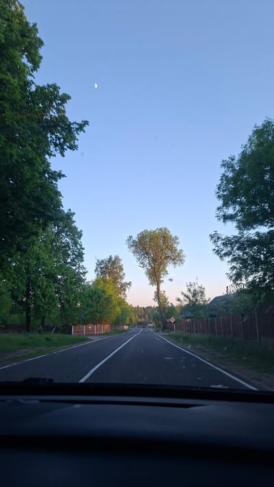 A photograph taken from inside a vehicle depicts a road landscape in Любанскі сельскі Савет, Belarus. The road is asphalt, with a single white line marking the lanes. Trees line both sides of the road, with a few wooden fences visible between the trees. The sky is light blue with a visible crescent moon. A yellow road sign is partially visible on the right side of the road. The vehicle's interior, including a dark grey section, is visible on the right side of the image.