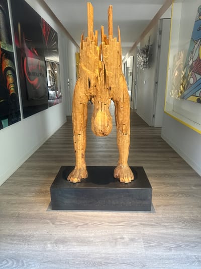 This is a photograph of a wooden sculpture of a person standing on a dark rectangular base. The sculpture is made of stacked, rough-hewn wooden pieces, giving it a primitive and abstract appearance. The figure is elongated, with clearly defined limbs and a head. The sculpture is positioned in a hallway or gallery space with a wooden floor. On the left wall, there are several framed artworks, including a brightly colored abstract piece at the top and a series of darker, graphic prints below. A metal art installation is visible on the ceiling in the background. The lighting suggests it is daytime, with natural light entering from an unseen source. The overall impression is one of a curated art exhibition. No text is clearly visible in the image.