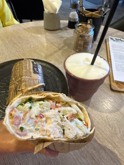 This is a close-up, eye-level shot of a table in a cafe, likely in Kirishi, Russia, given the provided context. The central subject is a half-eaten wrap, held in a person's hand. The wrap appears to be filled with chicken, vegetables like tomatoes and cucumbers, and a creamy dressing, all enclosed in a flatbread. A second, whole wrap, partially wrapped in newspaper-like paper, sits on a dark plate to the left. To the right, a mauve-colored cup contains a frothy beverage, likely a latte or cappuccino, with a black straw. In the background, a glass jar filled with sugar cubes and a metal holder with utensils are visible on the table, suggesting a casual dining setting. The overall lighting is soft and natural, creating a relaxed atmosphere.