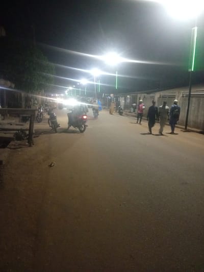 The image depicts a nighttime street scene in Tundun Wada West, Nigeria. The street is illuminated by streetlights, which cast a bright glow and create strong highlights and shadows. Several people are visible walking along the road, some in groups and others individually. Most of them are men, dressed in casual clothing, with some wearing traditional attire.

Several motorcycles are parked or moving on the street, suggesting this is a common mode of transportation. There are buildings and structures on either side of the road, including what appears to be a fence or wall along the right side. The overall setting is urban, with evidence of ongoing activity despite the late hour.

The time of day is clearly night due to the artificial lighting and darkness. There are no explicit weather cues, but the scene appears dry. The presence of motorcycles and people walking suggests a relatively active nighttime environment. The mood of the image is somewhat subdued, typical of nighttime scenes, with the focus being on the illumination and the figures navigating the darkened street. No visible text is present in the image.