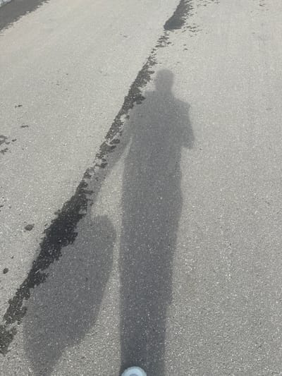 A shadow of a person is cast on an asphalt road. The shadow shows a person holding a suitcase. The road appears to be a street in the city of Snt Del'fin, Russia.