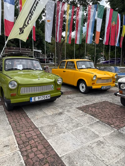 In Hadzhidimovo, Bulgaria, on a paved outdoor area with a brick pathway, a vintage car show is underway. The foreground features two classic Trabant cars. The one on the left is lime green, with a license plate that reads "E 9170 KH BG." It has a luggage rack on its roof with a brown vintage suitcase. The car on the right is bright yellow and displays a license plate that says "E 6424 BA." In the background, a line of national flags is displayed, fluttering in the breeze, with a wooded area behind them. Another car, possibly white, is partially visible on the far right. A purple car with writing on its side is parked further back, in front of some trees. People are visible inside and around the cars.