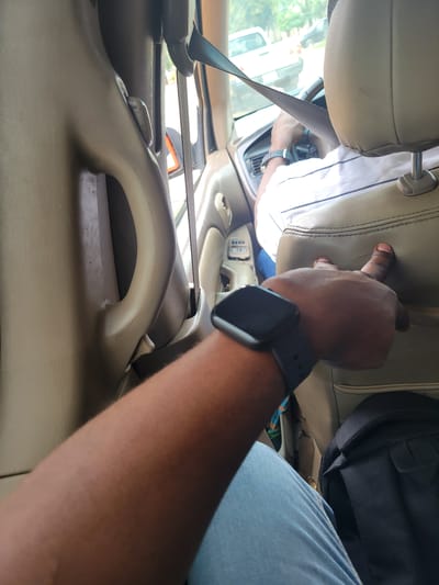A first-person perspective shot captures a person inside a vehicle, likely a car, in Katambe, Nigeria. The view is directed towards the front of the vehicle, with the driver's seat visible. A person in the driver's seat, wearing a white shirt with thin black stripes, is holding the steering wheel and appears to be driving. A seatbelt is fastened across their chest. The interior of the car is a light tan color, featuring a textured upholstery on the seats and door panels. A black smartwatch is visible on the left wrist of the person taking the photo. In the background, through the car window, a street scene is discernible, with a white car and trees suggesting an urban or suburban environment. The lighting indicates it is daytime.