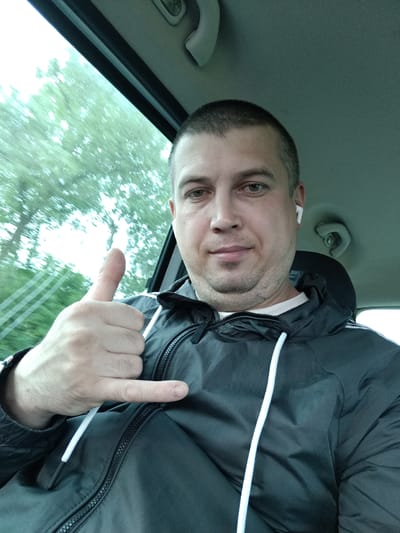 A man is taking a selfie in a car, giving a thumbs-up gesture. He is wearing a black hooded jacket with white drawstrings. A white wireless earbud is visible in his right ear. Trees and a slightly blurred landscape are visible through the car window, suggesting motion. The image appears to be taken during the daytime.