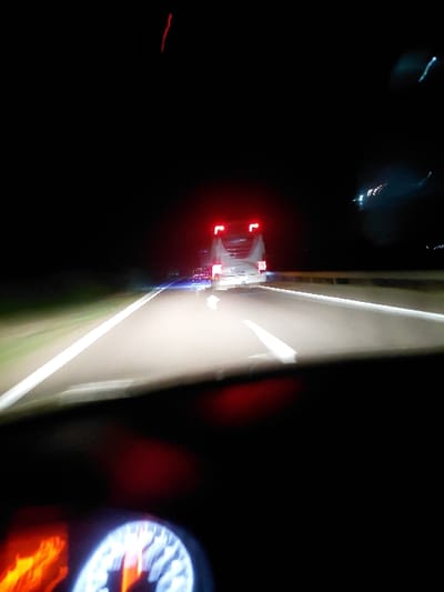 A nighttime scene blurred with motion depicts a vehicle ahead, its red taillights visible. An illuminated dashboard gauge sits in the foreground, possibly a speedometer. The image captures a blurry view of a road or highway, marked by white lines. The dark surroundings suggest a moving perspective from within a car, highlighting speed and darkness.