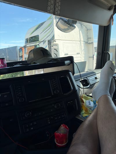 The image captures an interior view from the cabin of a large vehicle, likely a truck, possibly located in Parvenets, Bulgaria (though this cannot be definitively determined from the image). Prominently displayed is the control panel of the vehicle, featuring various buttons, screens, and controls. Resting on the dashboard are a pair of socked feet, indicating the driver is relaxed. A bag of "Maliva" snacks and a cup are also visible. Through the windshield, other trucks are seen on the road, suggesting the vehicle is in transit or parked in a truck stop or similar location. The sky is blue, indicating daylight.