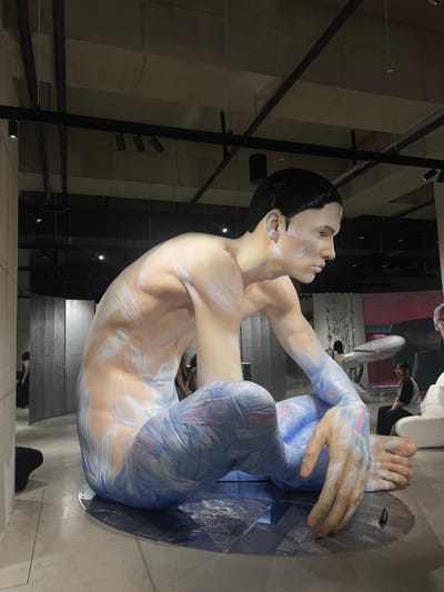 The image displays a large, seated male figure, appearing to be a mannequin or statue, in an indoor exhibition space located in 성동구, South Korea. The figure is positioned facing right, with its legs bent and crossed, and its arms resting on its knees. Its body is painted with visible brushstrokes of white, light blue, and hints of red/pink, giving it a textured appearance; white strokes are prominent on the torso and arms, while blue and red/pink tones cover the legs. The figure has dark hair and a pale complexion, resting on a dark, circular base on a light gray floor.
The surrounding environment is a modern interior with exposed concrete ceilings, black track lighting, and visible ductwork. To the left, a textured, silver-gray paneled wall is visible, with a partially obscured person in dark clothing near its edge. In the mid-ground and background to the right, various abstract white and light-colored furniture or sculptural elements are present. A person wearing a light gray shirt and dark pants is seated on a white, curved chair in the right background, facing away from the primary subject. Further back, a large dark panel exhibits a reddish-pink illumination. The lighting is generally subdued, with spotlights highlighting the main figure.