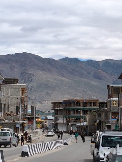 The image depicts a street scene in Padum, India, with a backdrop of imposing, barren mountains. The foreground features a paved road lined with black and white striped concrete barriers. On the road, several vehicles are visible, including a silver minivan on the left, a white SUV partially obscuring the view on the right, and a white car further down the street. People are walking along the road and sidewalks. Buildings line both sides of the street. On the left, a multi-story structure under construction with exposed concrete and scaffolding is prominent. Other buildings appear to be older, with varying architectural styles, some with visible construction elements or weathered facades. The sky is overcast with scattered clouds. The overall impression is of an active street in a town nestled amidst a rugged mountain landscape.