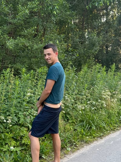 A young man in a teal t-shirt and navy shorts is standing in front of dense green foliage, with a paved path to his right. He is facing away from the camera and looking over his left shoulder with a slight smile. His right hand is obscured by his shorts. The background is dominated by lush green trees and smaller plants with white flowers. The lighting suggests it is daytime, possibly late afternoon.