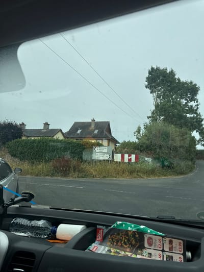 The image shows a view from inside a vehicle looking out at a rural intersection. The intersection features a white signpost with directional arrows and text indicating "km Ceanannas 9 KELLS R697" and "Cill Cheannain R697". There are also smaller signs indicating "L2630" and a red and white striped marker. In the background, two houses with tiled roofs are visible behind a dense green hedge. A mature tree with dark green foliage stands to the right of the intersection. The sky is overcast and appears light gray. The foreground shows the interior of the vehicle, including a water bottle, a roll of toilet paper, and some promotional flyers or magazines with prices displayed in Euros. A silver car is partially visible on the left side of the frame. The road surface is asphalt.