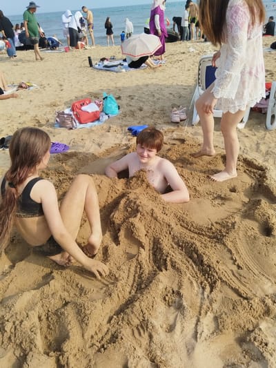 The image depicts a sunny day at a beach in Qinhuangdao, China, with the ocean visible in the background. The focus is on two children partially buried in the sand. On the left, a girl with long brown hair, wearing a black bikini top and bottoms, is sitting with her legs extended. On the right, a boy with short brown hair and a necklace is lying down, submerged in the sand up to his chest. Other beachgoers and various objects like bags, shoes, and beach umbrellas are scattered around the scene. The overall atmosphere appears relaxed and playful, suggesting a family outing or a day of recreation at the beach. The time of day is likely midday or early afternoon, given the bright sunlight and the shadows cast on the sand.