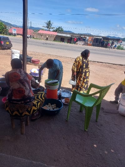 An outdoor scene in Kimamba, Tanzania, depicts a roadside food preparation and vending area under a clear sky with scattered clouds. In the foreground, a person is seated on a small wooden stool, their back to the camera, wearing a floral-patterned top and a geometric-patterned skirt. To their right, a person in a light blue garment is bent over, actively stirring or preparing food in a white basin placed on a low surface. Adjacent to this activity, a person wearing a long yellow and black patterned dress and a dark head covering stands upright, holding a white container.
Surrounding these individuals are various items: multiple plastic basins (red, purple, blue) containing food items, an orange and green thermos, metal trays, and small plates. An empty green plastic chair is positioned to the right of the central activity. The ground is composed of dirt and gravel.
In the background, a paved road stretches across the image, with a dark-colored SUV parked on the left shoulder. Along the opposite side of the road, several one-story buildings with reddish-brown roofs are visible, appearing to house small shops or stalls. Distant hills or mountains frame the horizon under the bright sky. Several indistinct figures are visible near the roadside buildings and vehicles, indicating general street activity.