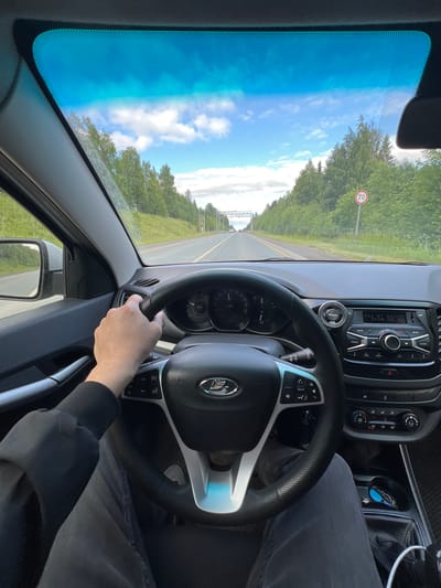 The image shows the interior of a car, looking forward through the windshield. A person's hands are on the steering wheel. The car is driving on a paved road with trees and greenery on either side. There is a speed limit sign indicating 70. The sky is blue with some clouds. The dashboard and radio of the car are visible. The location is identified as Verkhne-Pozim', Russia.