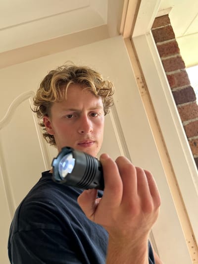 A male subject, with blonde, wavy hair and a dark t-shirt, is depicted indoors in Thirroul, Australia. He holds an illuminated black cylindrical flashlight in his right hand, angled forward. The subject maintains direct eye contact with the viewer, exhibiting a serious expression. To his left, behind him, is an off-white panel door and its frame. To his right, an opening reveals a section of an exterior red-brick wall and a light-colored eave above it. The door frame adjacent to the brickwork shows minor surface wear.