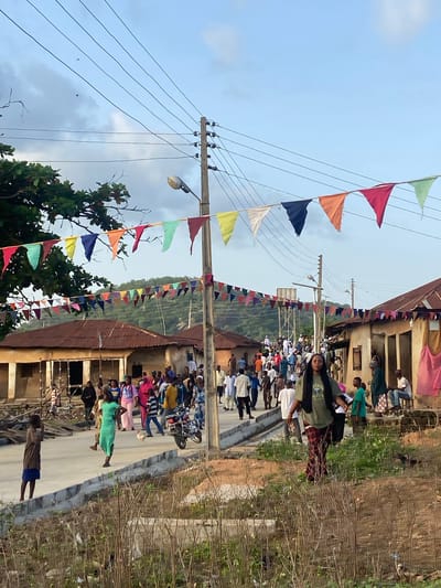 The image shows a street scene in Ozuri, Nigeria. A crowd of people are gathered in the street, and some are kneeling. Flags are strung across the street. Power lines and utility poles are visible. There are buildings on either side of the street. A woman with long hair is standing on the right side of the street. Vegetation, including trees and shrubs, are also present.