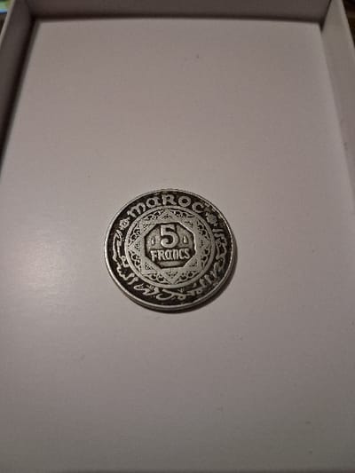 A Moroccan 5 Francs coin is displayed on a white surface. The coin is silver in color and features intricate geometric patterns. The number "5" is prominently displayed, along with the word "FRANCS". The outer ring of the coin contains the inscription "MAROC" and Arabic script. The coin appears to be aged and slightly tarnished.