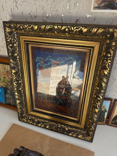 The scene depicts an indoor display of objects within a structure located in Matveevskoe, Russia. A large, ornate framed artwork is centrally positioned, featuring a dark brown frame with extensive gold scrollwork and a secondary inner gold frame. The artwork itself, a mosaic or beaded piece, displays two dark, stylized figures on a textured dark base against a starry blue background. Reflections on the glass cover obstruct parts of the artwork, revealing reversed text fragments including "KROH" and "LONDON".
To the left of the main artwork, a smaller wooden-framed photograph shows two young children, one wearing a green top, the other a lighter top, both smiling. A blue object is partially visible next to this photo. To the right, two small religious icons stand vertically; one portrays a female saint in a green robe. The background wall exhibits a textured, silvery-grey, distressed finish. In the immediate foreground, a light brown surface supports a dark, irregularly shaped, textured object. No individuals are present in the immediate scene.