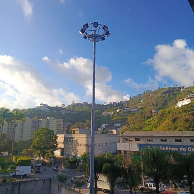 The image is a daytime, eye-level, wide shot of a residential and commercial area in Carrizal, Venezuela. In the foreground, a tall, metallic lighting pole with multiple floodlights is positioned centrally. Behind it, a street level view shows a building with the text "CENTRO COMERCIAL LOS ALTOS" visible, and another building with the text "COLINAS DE CA" in blue letters. There are several vehicles on the road, including a white truck, a white pickup truck, a silver SUV, and other cars. The midground features dense green trees and palm trees, as well as more residential buildings, including a tall, light green apartment complex. The background displays a hillside covered in lush green vegetation and numerous houses and buildings, indicating a densely populated urban environment on a slope. The sky is blue with scattered white clouds. The lighting suggests it is either early morning or late afternoon. There are a few individuals visible walking on the street and near the buildings, but their features are not discernible.