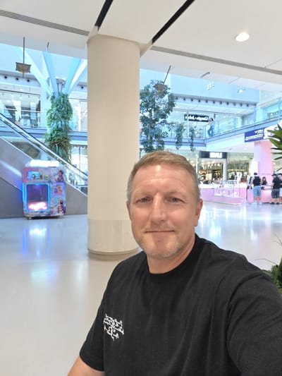 The image is a selfie taken inside a brightly lit, modern shopping mall. The man in the foreground, who is the subject of the selfie, is wearing a black t-shirt with a white graphic on the left sleeve. He has short, light-colored hair and a light beard. The background shows a wide-open mall interior with escalators, shops, and decorative plants. To the left of the man, an escalator leads upwards, and a brightly lit vending machine with cartoon graphics is visible beside it. Further in the background, shops with glass fronts and various displays are apparent, and people are seen walking and browsing. The overall impression is of a clean, spacious, and contemporary commercial environment. The location of the image is not specified as Nola, Italy.