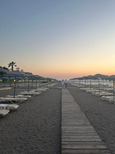 Two people, dressed in light-colored clothing, walk away from the camera on a wooden boardwalk bordering a dark sand beach. They are walking towards a soft gradient of sunset colors in the sky, transitioning from pale yellow near the horizon to a soft blue overhead. The beach is lined with numerous empty lounge chairs and closed umbrellas, suggesting a quiet time of day, likely early evening. The sea is visible in the distance, calm and reflecting the sky's colors. Palm trees are sparsely scattered along the edge of the beach, further indicating a warm, possibly tropical or Mediterranean, location. The overall atmosphere is serene and peaceful. No visible text is present in the image. The scene is set on a beach in Rhodes, Greece, during sunset.