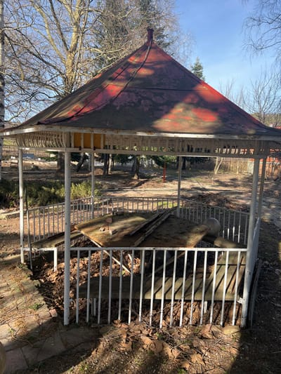 An abandoned hexagonal pavilion stands in an unkempt outdoor area in Panichkovo, Bulgaria. The pavilion features a conical roof, heavily discolored and peeling from an original red finish, showing patches of pinkish-purple and green moss. The supporting structure consists of white metal posts and railings, exhibiting significant rust and chipped paint. Inside, the ground is covered in dry leaves and debris. Slatted wooden benches line the interior perimeter; some sections are broken. A large, flat wooden panel lies diagonally across two benches, with two reddish bricks resting on it. Another white, spherical object is visible on a bench section to the right. The surrounding environment consists of bare soil, dry leaves, and leafless deciduous trees under a clear, bright sky. No individuals are present in the scene.