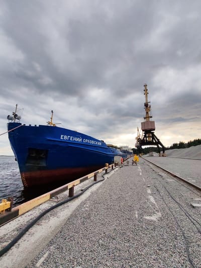 The image depicts a blue cargo ship named "Евгений Орловский" docked at a port in Shal'skii, Russia. The ship is positioned alongside a concrete pier covered in gravel. In the background, industrial cranes stand tall, indicating port operations. Several piles of gravel are visible on the right side of the image, suggesting the cargo being handled. The sky is overcast with dark, heavy clouds. A few individuals are present on the pier, possibly port workers. The scene conveys a sense of industrial activity in a cloudy, maritime environment.