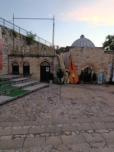 The image shows the exterior of the Nisville Jazz Museum, located in the municipality of Crveni Krst, Serbia. In the foreground, a cobblestone courtyard leads up to a set of stairs with green carpeted risers, each displaying the text "NISVILLE JAZZ MUSEUM" in black. Above the stairs, the stone facade of the museum building is visible, with arched doorways and windows. Large, stylized cutouts of a saxophone and a cello stand outside the entrance to the right. The museum's name, "JAZZ MUSEUM," is displayed in large, weathered concrete letters above the building. The background features a stone wall, a railing, and a hint of a dome atop another structure. The sky is clear with a crescent moon visible. There are no people clearly visible in the scene, only silhouettes in the distance. The environment appears to be an outdoor public space.