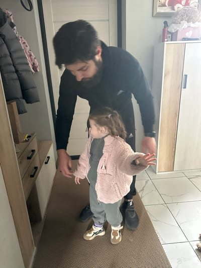 The image captures an intimate indoor moment between an adult man and a young child, likely in a hallway or entryway. The original orientation of the image is rotated 90 degrees clockwise.
The man, with dark hair and a beard, is dressed in a black long-sleeved shirt or hoodie and black pants, wearing black sneakers, one of which shows a faint Adidas logo. He is leaning down, looking towards the child with an attentive gaze.
The young girl has light brown hair tied back with a purple hair tie. She is wearing a soft, fluffy pink jacket, grey sweatpants, and light-colored sneakers with dark accents. She stands facing the man, with one hand raised, palm open, in a gesture that could be playful, a greeting, or reaching out.
The setting features white tiled flooring with a subtle grey veining pattern, partially covered by a brown, textured, perforated floor mat. Various storage units are visible in the background: an upper wall-mounted cabinet with a wooden drawer, slightly open to reveal a framed picture, white fabric, and a red object. In the lower part of the image, there's another wooden cabinet with black handles, its top drawer ajar, revealing a yellow item inside. Stacked grey padded jackets and a pink patterned item (possibly a blanket or another jacket) are also visible on the floor, suggesting a practical, lived-in space. The scene conveys a warm, everyday interaction between the two individuals.