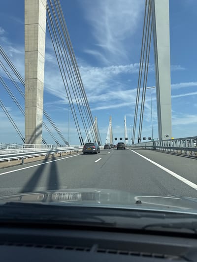 The image captures a view from a vehicle driving on a multi-lane cable-stayed bridge, likely in the vicinity of Andelst, Netherlands, given the distinctive architecture of the bridge towers. The sky above is a clear blue with scattered white clouds. The bridge features prominent concrete and white pylons from which numerous cables extend to support the roadway. Several cars are visible on the road, all traveling in the same direction as the viewer's vehicle. The road surface is asphalt with white lane markings. The foreground shows the dashboard and windshield of the vehicle, with reflections of the bridge structure and sky visible on the windshield. The environment is characterized by the vastness of the bridge structure against the open sky, suggesting a location over water or a significant geographical feature.