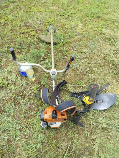 A top-down, high-angle, medium shot shows a disassembled string trimmer and related accessories lying on a patch of overgrown green grass with some sparse yellow wildflowers. The string trimmer consists of an orange and white motor unit with a black strap and padding. To the left of the motor is a silver shaft with black and orange handles and a green spool of string. A clear plastic bottle with a yellow liquid is situated next to the spool. To the right of the motor, a black and yellow shield-like attachment with straps is positioned on the ground. The scene is located outdoors, in the city of Гожскі сельскі Савет, Belarus.