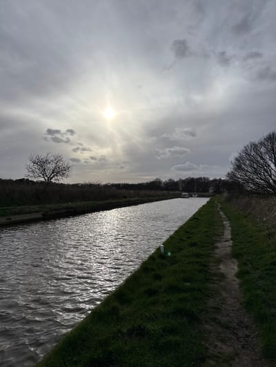 This image depicts a straight, narrow waterway, likely a canal or river, under a partially cloudy, grey sky in North Norfolk, United Kingdom. The sun is a bright, radiating point in the upper left quadrant, creating a strong lens flare and a prominent, shimmering reflection path across the dark, rippled water.

The waterway's left bank features a low, dark edge, possibly concrete, behind which dense, dry reeds and bare scrubland extend into the distance. A solitary, bare deciduous tree with a wide, gnarled canopy stands prominently on this left bank in the mid-distance. The right bank consists of a strip of green grass, approximately two meters wide, alongside which runs a well-trodden, unpaved dirt path, roughly one meter in width. Two small, white cylindrical posts are visible on the grassy verge between the path and the water, spaced along the bank.

In the far distance on the waterway, two white leisure boats are visible, appearing stationary or slowly moving away. The horizon beyond the waterway and banks is marked by a line of distant, bare trees. The overall lighting indicates backlighting from the low sun, creating strong contrast and shadowing parts of the foreground path and banks. No individuals or animals are present in the scene.