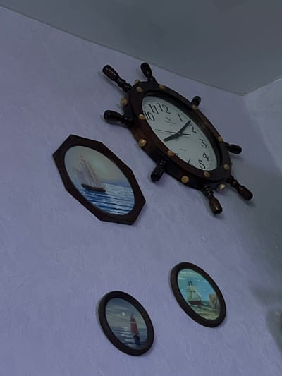 The media file shows a wall decorated with nautical-themed items. The primary subject is a ship's wheel clock, made of dark wood with brass accents and a white face displaying Roman numerals. The clock hands indicate it is approximately 9:05. To the right of the clock, there are two oval-shaped framed pictures. The upper picture appears to depict a ship sailing on the water under a bright sky. The lower picture shows a lighthouse on a rocky outcrop with a calm sea. The wall has a textured, pale purple finish. The scene suggests a home or interior space, likely in Bely Klyuch, Russia, given the location context. No people are visible in the frame. The lighting suggests it is daytime.