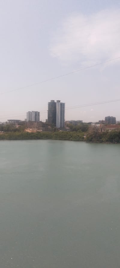 The image presents an expansive view of Tudor ward, Kenya, observed from an elevated perspective across a body of water. The foreground features a calm, light blue-green water surface with minor textural variations. A dense strip of green vegetation, consistent with mangroves, borders the water and separates it from the urban landmass. The mid-ground and background are occupied by an urban skyline dominated by numerous multi-story buildings. Two prominent high-rise structures stand in the center-right; one is a light-colored, seemingly completed or near-completed residential building with visible balconies, while the adjacent structure to its left appears darker and is actively under construction, partially obscured by scaffolding. Additional multi-story buildings, some complete and others exhibiting exposed concrete frameworks indicative of ongoing construction, extend across the visible cityscape. The sky, occupying the upper third of the frame, is hazy and light blue, with faint, diffuse white cloud formations. Two thin, dark horizontal lines, likely utility wires, traverse the upper section of the image, above the distant buildings. No individuals or discernible activities are observed within the scene.