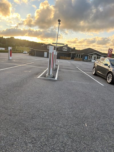 The image depicts an outdoor parking lot at sunset, likely in Hogstorp, Sweden, judging by the presence of a "OKQ8" gas station sign and what appears to be "STUS" visible on a building. The sky is filled with dramatic, golden-hued clouds illuminated by the setting sun. In the foreground, asphalt parking spaces are marked with white lines. Several Tesla Supercharger stations, white with red accents, are prominently positioned. A dark-colored Tesla Model X is parked in the right side of the frame, partially visible. In the middle ground, a low-rise building with a dark facade, a pitched roof, and several windows is visible. This building appears to be a commercial establishment, possibly a restaurant or convenience store, indicated by the "OKQ8" signage and a smaller sign that may read "RESTAURANT." Further in the background, a white car is seen parked in a lot, and beyond that, a wooded hillside under the twilight sky. The overall scene suggests a functional, publicly accessible area designed for vehicle charging and services.