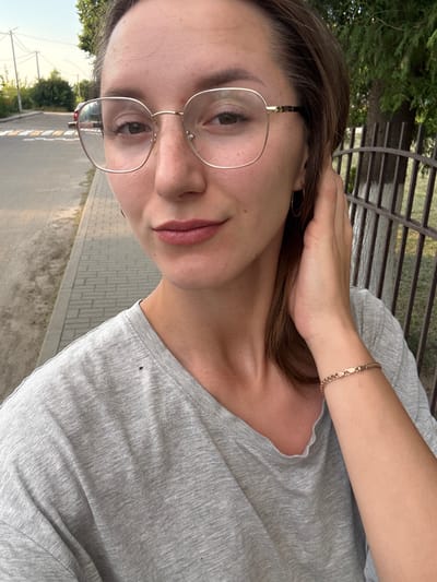 This is a vertical, close-up, selfie-style photograph of a young woman with brown hair and glasses. She is wearing a light gray t-shirt. Her left hand is raised to her head, touching her hair. A thin gold bracelet is visible on her right wrist. She is wearing light-rimmed glasses and has light pink lipstick on her lips. The background shows a blurry outdoor scene with trees, a fence, and a paved walkway. The lighting suggests it is daytime, likely late afternoon or early evening, given the warm tones. The image has a casual and relaxed feel, possibly taken while sitting or standing outdoors. The location is identified as Zhodzina, Belarus. There is no visible text in the image.