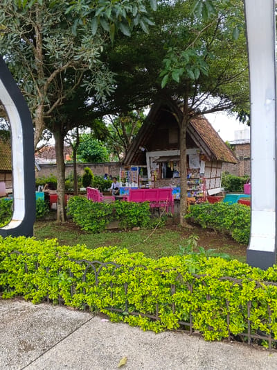 This image depicts an outdoor recreational area in thành phố Biên Hòa, Vietnam. The scene is dominated by a small, traditional-style kiosk situated towards the center-right, featuring a brown tiled roof, light-colored walls with dark horizontal and vertical framing, and an open service window. The kiosk displays various packaged goods and snacks on shelves and racks. Surrounding the kiosk are several brightly colored benches, predominantly pink, with some red and blue seating visible further back.
Multiple mature trees with green foliage provide shade over the area. The ground is covered with grass and interspersed with manicured bright green shrubs, particularly forming a low hedge in the foreground, bordered by a dark metal fence. Beyond this fence, a speckled concrete or asphalt path is visible.
Several individuals are present. One male subject, wearing a dark shirt and light pants, is seated on a pink bench to the left of the kiosk, appearing to interact with a personal device. Another person is partially visible seated on a red bench further to the left. Other indistinct figures are visible in the mid-ground, suggesting more people are present within the shaded area. A brick wall runs along the background, partially obscured by trees. On the far left, a segment of a large, black and white curved structure is visible, and on the far right, a vertical white post. The lighting suggests daytime conditions.