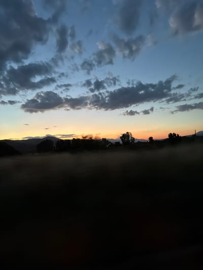 The image depicts a twilight scene in a rural area, possibly on the outskirts of Damianitsa, Bulgaria. The sky is a gradient of deep blue at the top, transitioning to lighter blues and then to a band of warm orange and yellow near the horizon, indicating either sunrise or sunset. Dark, scattered clouds drift across the sky, adding texture and depth. Below the sky, a dark silhouette of a treeline is visible against the illuminated horizon, with some faint lights suggesting buildings in the distance. The foreground is dominated by a dark, blurry expanse, likely a field or open ground, with a wispy layer of mist or fog obscuring the details. The overall impression is one of quiet and natural beauty, characteristic of a rural landscape at the transitional hours of the day.