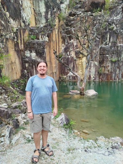 A man with shoulder-length brown hair and a beard stands smiling in front of a large, rocky cliff face and a body of clear green water. He is wearing a blue t-shirt, khaki cargo shorts, and gray hiking sandals. Behind him, the cliff is a mix of light and dark rock layers with streaks of orange and brown. A gnarled, leafless tree grows out of the water near the center of the image, with its branches reaching towards the cliff. Several large rocks are visible in the water around the tree. The ground in front of the man is covered with small stones and some sparse green vegetation. The water appears shallow near the edge of the shore, with rocks and sand visible beneath the surface. The overall scene suggests an outdoor location, possibly a quarry or a natural rock formation. The image does not provide information about its specific location within Lán Tranh, Vietnam.