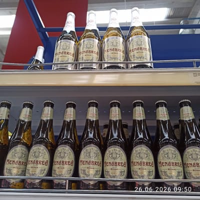 This image displays a close-up view of a refrigerator shelf stocked with bottles of Menabrea beer. The bottles are predominantly dark brown glass with light-colored labels featuring intricate illustrations and Italian text. The labels prominently feature the brand name "MENABREA" in large, stylized lettering, along with descriptions like "BIONDA," "PREMIUM," and "LAGER." Some labels also indicate "La 150," suggesting a 150th anniversary, and mention "CASA FONDATA NEL 1846" (House founded in 1846) and "BIELLA-ITALIA." The bottles are arranged in multiple rows on metal shelving within the refrigerator. A bright light source is illuminating the scene from the left, creating reflections on the glass bottles. The background shows a glimpse of a blue metal structure and a cream-colored vertical panel with some text, possibly a price tag or product information. On the far right of the image, a timestamp "26.06.2026 09:50" is overlaid in white text. The overall scene appears to be a retail environment, likely a supermarket or liquor store, given the presentation of the bottled beverages. The focus is on the product, with no people visible.