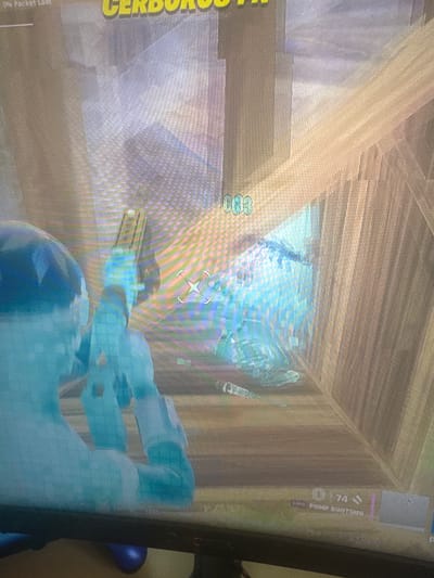 The image shows a first-person perspective of a video game, likely Fortnite, as indicated by the user interface elements. The player character, rendered in a translucent blue, is holding a weapon, possibly a shotgun, which is indicated by the "EPIC PUMP SHOTGUN" text at the bottom right. The game environment appears to be a building with wooden walls and floors. There are also various UI elements visible, including "0% Packet Loss" in the top left corner, "CERBERUS III" text, and a numerical display "003" in the upper center. The player is aiming at a scattered collection of items on the floor, which include what appear to be weapon parts or ammunition. The lighting suggests an indoor, potentially low-light environment. There is no discernible location within the city of Bezden, Bulgaria, as this is a digital representation within a video game.