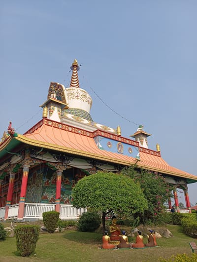 The image shows a colorful Buddhist temple in Lumbini Sanskritik, Nepal. The temple features an orange roof, intricate golden detailing, and vibrant murals on its exterior walls. A white stupa-like structure with a golden spire rises from the top of the building. In the foreground, there is a landscaped area with a small tree under which a group of statues depicting monks are seated in front of a Buddha statue. The sky above is clear and blue.