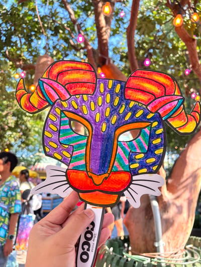 A person's right hand holds a vibrantly colored paper mask on a white stick, centered in the foreground. The mask depicts a stylized animal face, resembling an alebrije from Disney/Pixar's *Coco*, with large horns, a prominent snout, and eye cutouts. It is colored with markers in a patchwork style: the forehead and cheeks are purple with yellow oval spots; the horns are striped red, orange, and yellow; the areas around the eyes feature diagonal stripes of green, blue, and pink; and the snout/mouth is orange and red. White paper cutouts form whiskers or decorative fringe. The supporting stick bears the "COCO Disney Pixar" logo printed vertically in black. The background is out-of-focus, showing a sunny outdoor environment with green tree foliage and brown trunks. Numerous string lights with yellow, pink, and orange bulbs are visible strung among the trees. Blurred figures of other individuals are present in the background, one person on the left wearing a colorful, patterned shirt. This scene is located in Anaheim, California, United States, specifically within the Disneyland Resort, likely Disney California Adventure Park.