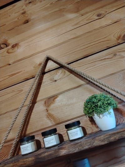 A close-up, eye-level shot shows a rustic wooden wall with a shelf attached. A thick, beige rope is suspended from an unseen point above, creating a triangular shape with two strands hanging down towards the shelf. On the shelf, to the left, are three small, clear glass jars with black lids, each with a label. The leftmost label appears to read "имбирный пряник," and the middle label reads "Рождественский." The rightmost jar has a label that says "Шампанское и клубника." To the right of the jars, a small, potted green plant with dense foliage sits in a white ceramic pot. The overall environment suggests an indoor, possibly home decor setting, in a location such as Vostochnaya, Russia, given the context of the request.