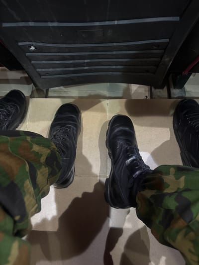 The image is a first-person perspective of someone wearing camouflage pants and black combat boots. The boots are laced and appear to be in good condition. The person is sitting or standing on a tiled floor, with the legs and boots prominent in the foreground. In the background, there is a black, ridged surface, possibly a seat or a piece of furniture. The lighting suggests an indoor or covered outdoor environment. There are no identifiable landmarks or features that would indicate a specific location in Birnin Yero, Nigeria, or any other city. The scene is devoid of other people or any contextual elements that would provide further information about the setting or the activity taking place.