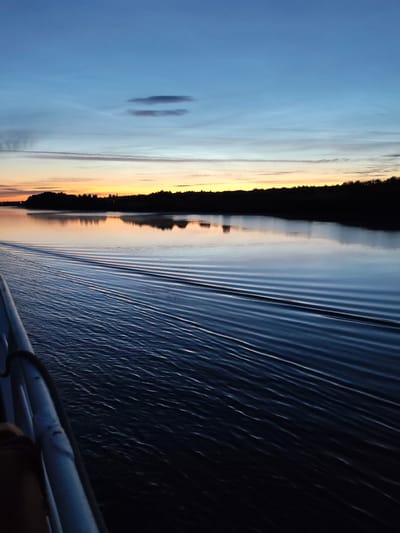 The image depicts a serene twilight scene on a body of water, likely a river or lake, with a faint sunrise or sunset glow on the horizon. The sky transitions from a deep blue at the top to lighter shades of blue and a warm orange and yellow band near the horizon, punctuated by a few wispy clouds. Silhouetted against the colorful sky is a dense line of trees forming the distant shoreline.
The foreground is dominated by the dark blue water, showing distinct ripples and waves created by movement, suggesting a boat or vessel is present and moving. A portion of a white railing from the vessel is visible on the left side of the frame, partially obscuring a person's arm. The overall impression is one of peacefulness and tranquility at the transition between day and night.
The location is specified as Matveevskoe, Russia. This setting suggests a natural landscape, possibly a rural or semi-rural area, consistent with the depiction of a wide expanse of water bordered by a forest. The time of day and the calm water further contribute to the peaceful atmosphere.